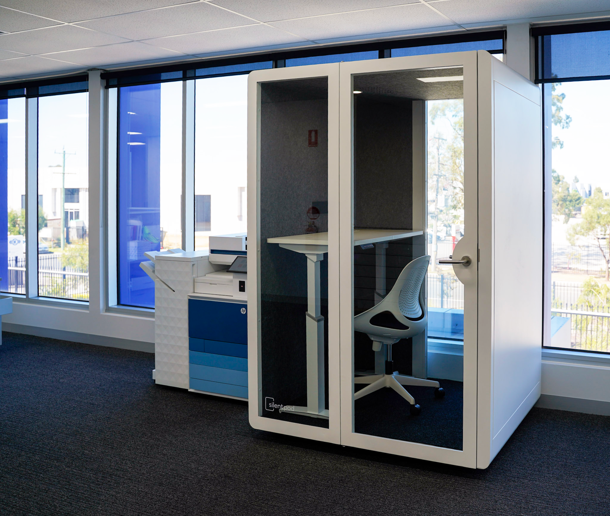 Side view of a white meeting pod with glass panels in a modern office near a blue and white printer station.