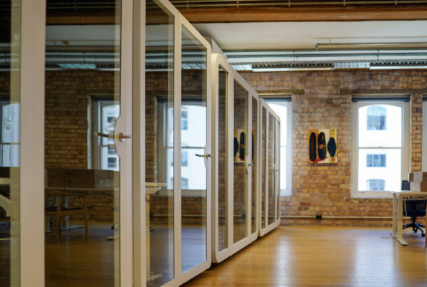 A row of modern soundproof office pods with glass doors, set inside a warm brick-walled coworking space with natural light and wooden floors.