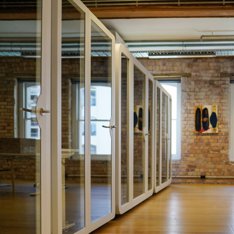 A row of modern soundproof office pods with glass doors, set inside a warm brick-walled coworking space with natural light and wooden floors.